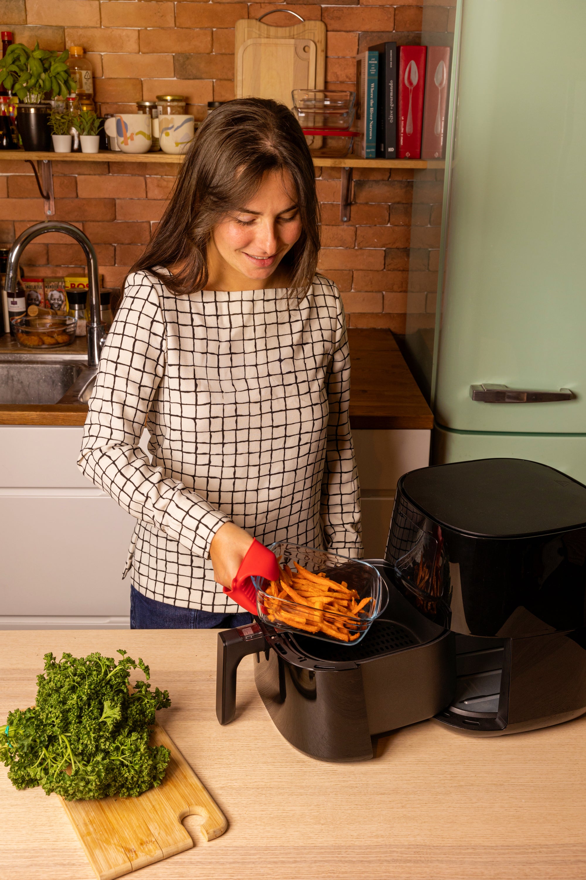Woman using an air fryer in a kitchen using an ÔCuisine square Air Fryer roaster with a refrigerator and wooden shelves in the background.