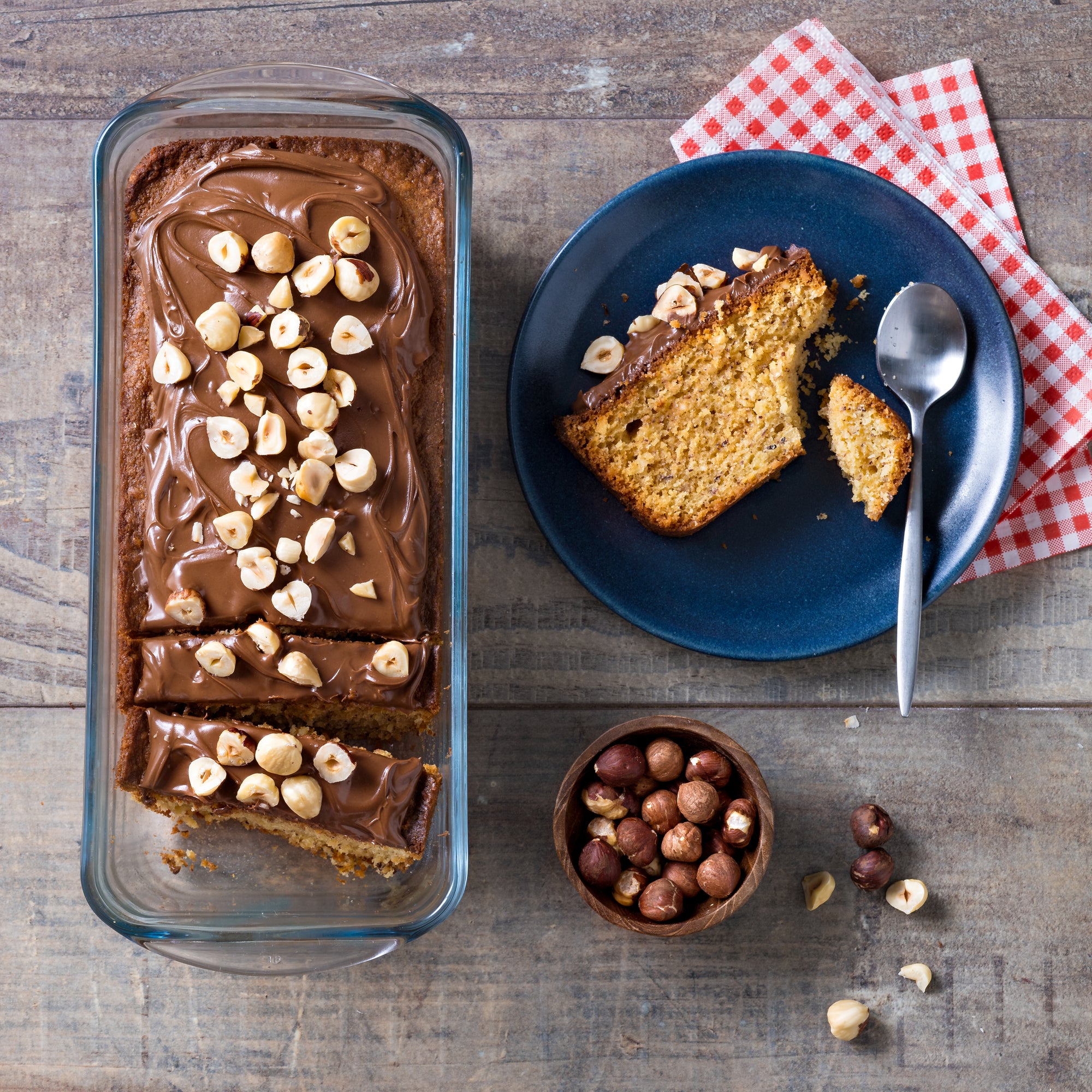 Loaf of cake with chocolate frosting and nuts in an ÔCuisine Loaf dish, served on a blue plate with a bowl of nuts and a spoon.
