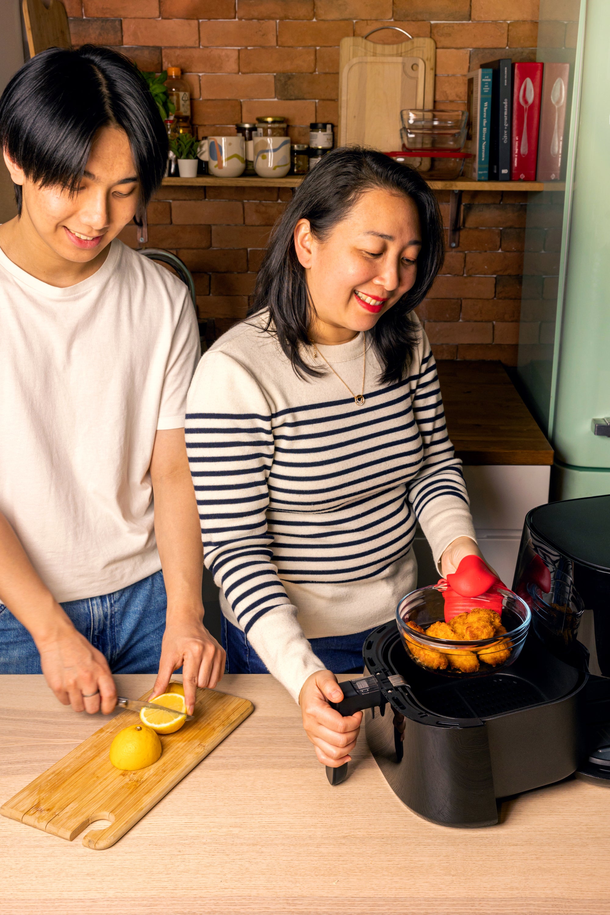 Two people cooking together in a kitchen, with one person cutting lemons on a wooden board and the other using a deep fryer.