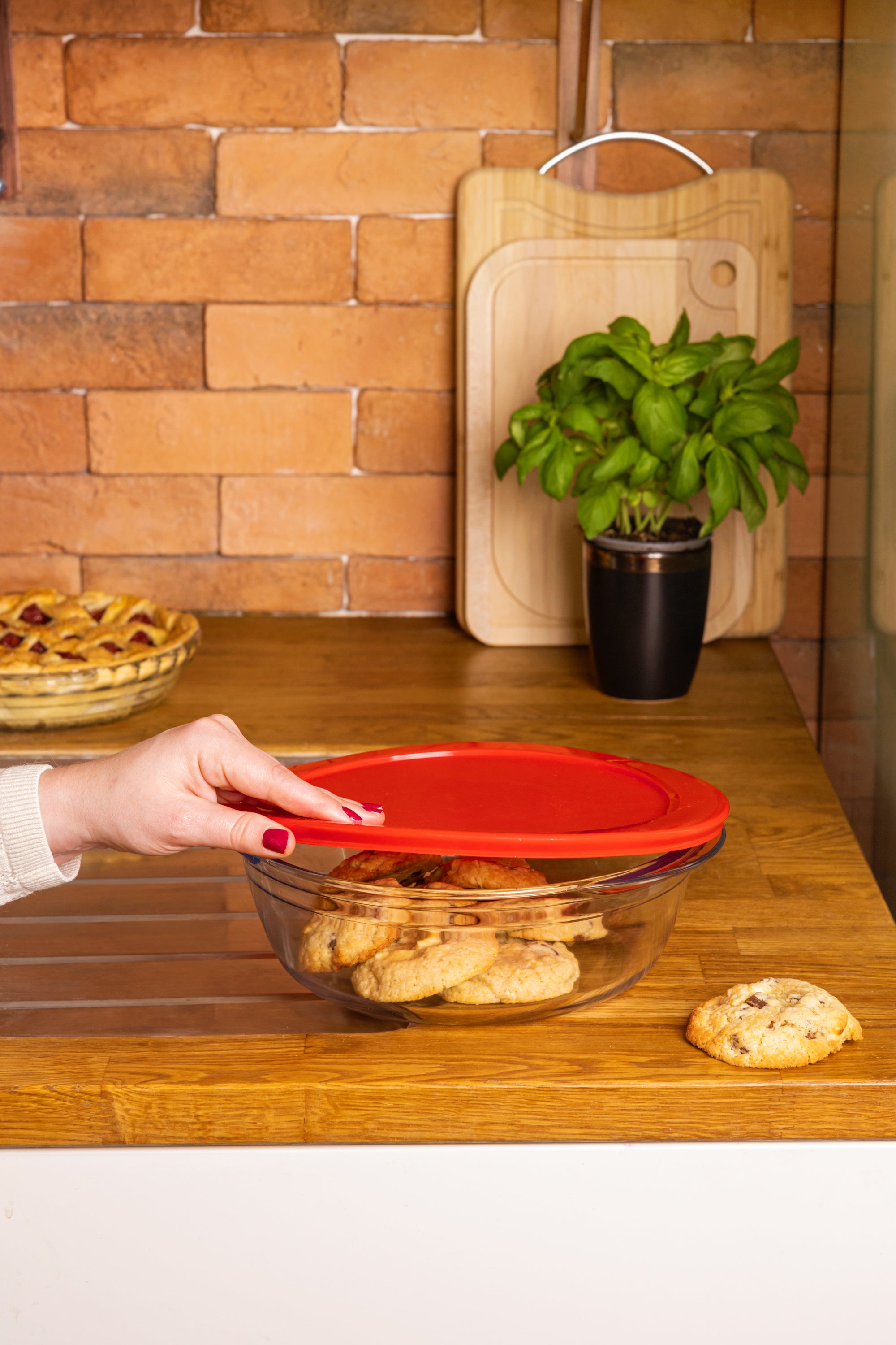 Person closing a glass container with a red lid on a wooden countertop, with a plant and cutting board in the background.