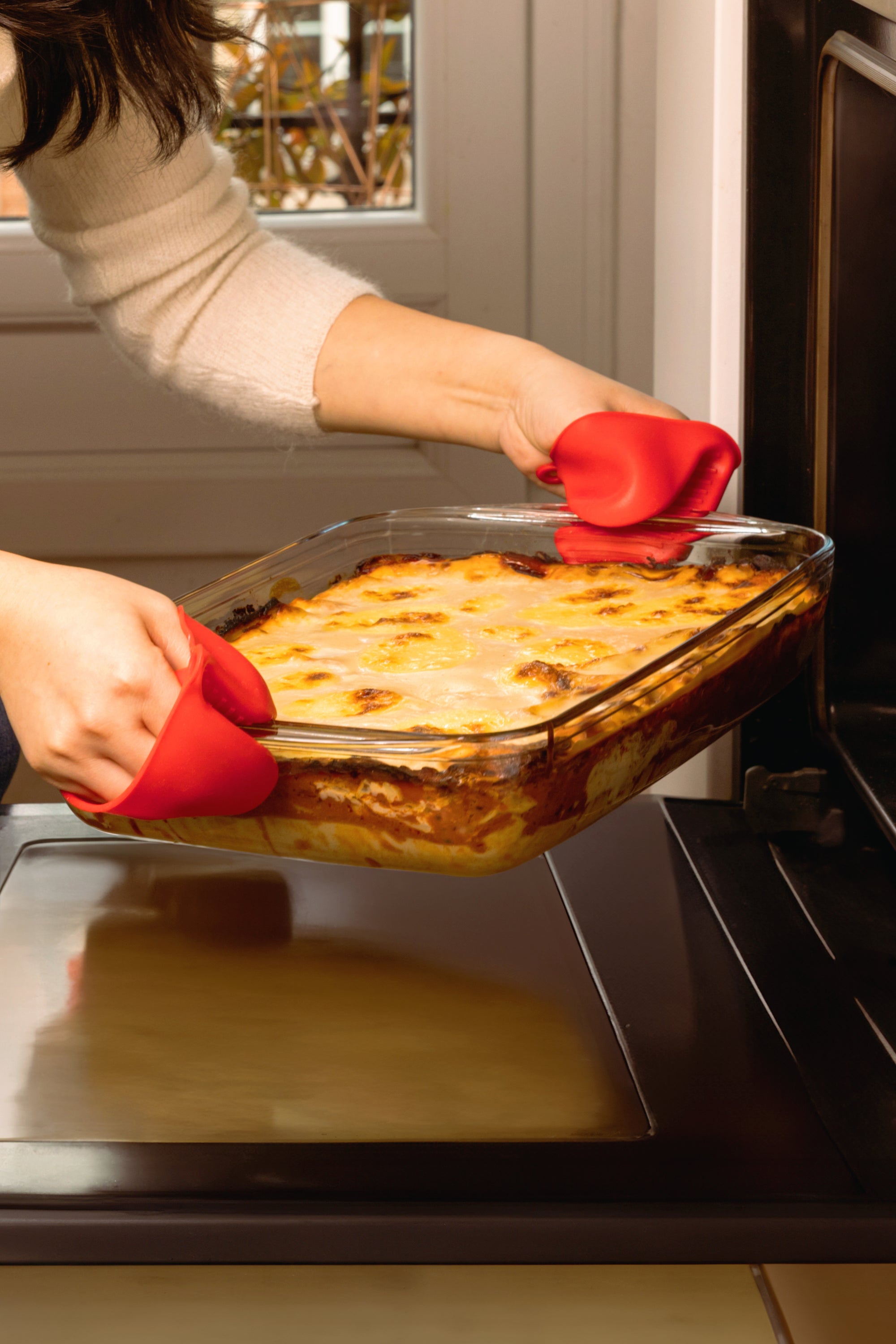 Person removing a dish of lasagna from an oven with red oven mitts.